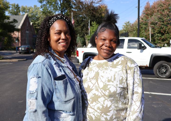Two women stand in a parking lot smiling at the camera, including the woman who is featured in the story and her case worker. Both women are dressed casually, after attending the Safe Sleep Summit.